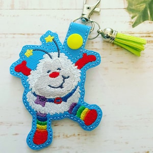 May include: A novelty keychain with a cartoon character design. The character has a blue outline, white body, and rainbow details. It features a silver clasp and a lime green tassel. The character has a smiling face and yellow stars.