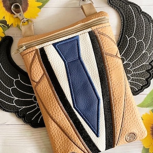 May include: A tan crossbody bag with a blue tie design, black and white accents, and a zip closure. The bag has a detachable strap and is accompanied by two black wing-shaped patches. The bag is approximately 20 cm wide and 25 cm tall.