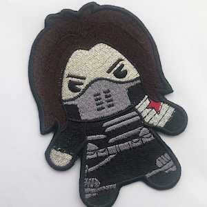 May include: Embroidered patch of a cartoon character wearing a black mask and a red star on their arm. The character has brown hair and is wearing a black and grey outfit.
