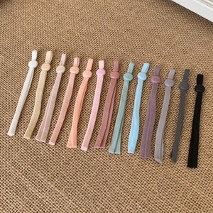 May include: A set of 14 colourful elastic cord mask extenders with round plastic stoppers. The extenders are in various shades of pink, blue, grey, and black.