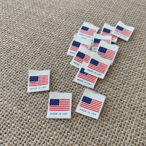 May include: A group of small, white fabric labels with an American flag and the text "Made in USA" printed on them. The labels are arranged on a brown burlap surface.