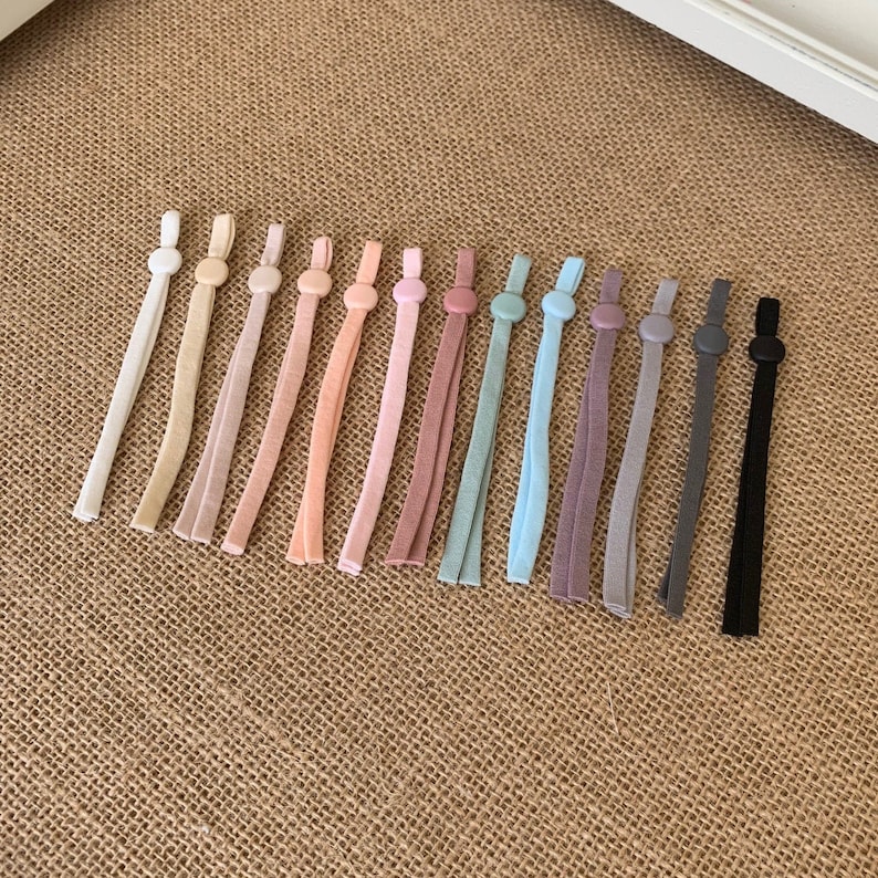 May include: A set of 14 colourful elastic cord mask extenders with round plastic stoppers. The extenders are in various shades of pink, blue, grey, and black.