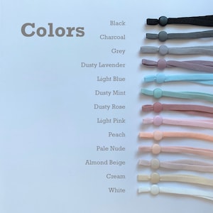May include: A range of colours of elastic cord with a button on the end. The colours are black, charcoal, grey, dusty lavender, light blue, dusty mint, dusty rose, light pink, peach, pale nude, almond beige, cream, and white.