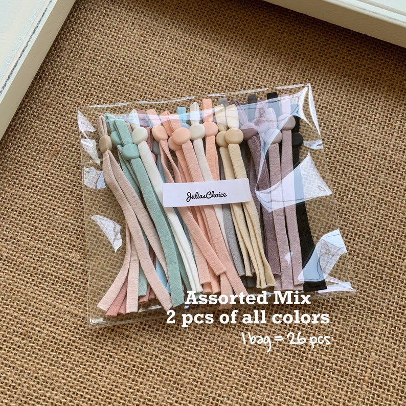 May include: A clear plastic bag containing 26 assorted colours of elastic mask straps. The straps are made from soft, stretchy fabric and have a small button on each end. The bag has a label that reads "JuliasChoice" and "Assorted Mix 2 pcs of all colours 1 bag = 26 pcs".