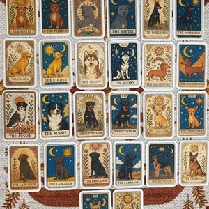 May include: A collection of tarot cards featuring various dog breeds. Each card has a unique illustration of a dog, with titles like "The Pittie," "The Golden," and "The Husky." The cards have a vintage aesthetic with celestial and nature-inspired designs.