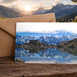 May include: A greeting card depicting a mountain scene with a building reflected in a lake. A brown envelope sits beside the card on a wooden surface. The background shows mountains and a sunset, evoking a sense of tranquility.