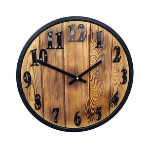 May include: Circular wall clock with a rustic design. The clock face is made of wood with a natural grain pattern. Black numerals and hands contrast against the light brown wood. The clock is framed with a black border.