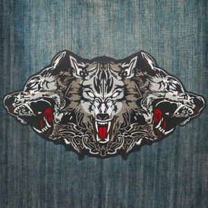 May include: Embroidered patch featuring a three-headed wolf design. The central wolf has a white face with a snarling expression and red mouth. The other two wolves have black faces with red mouths. The patch is on a blue denim background.