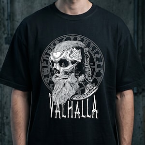 Könnte beinhalten: Ein schwarzes Kurzarm-T-Shirt mit einem weißen Grafikdruck eines bärtigen Schädels mit geflochtener Frisur. Das Design enthält ein Valknut-Symbol, eine kreisförmige Runenumrandung und das Wort VALHALLA in großen weißen Großbuchstaben.