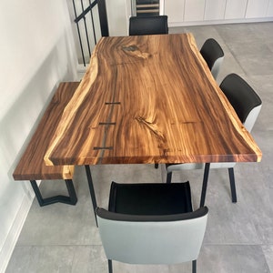 May include: A live edge wooden dining table with a matching bench and four chairs. The table and bench have a natural wood grain finish. The chairs have gray backs and black seats. The table is supported by black metal legs.