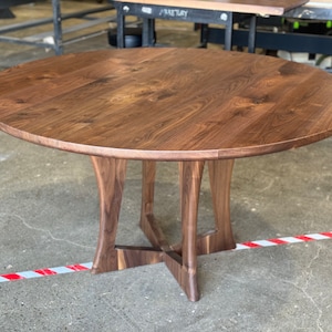 May include: A round wooden dining table with a dark brown walnut finish. The table has a unique, curved base with a cross-shaped support.