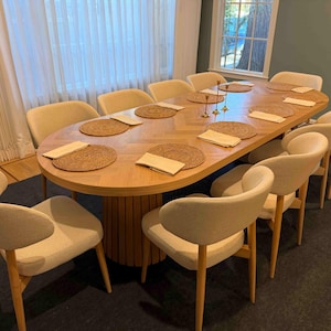 May include: A large, oval wooden dining table with a light wood finish, set for a meal. Ten cream-colored upholstered chairs surround the table. Woven placemats and folded napkins are arranged on the table. The room has a dark rug and a window.