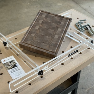 May include: A clear acrylic jig for routing a juice groove into a wooden cutting board. The jig is adjustable and has a clamp to secure the board in place. The jig is on a pegboard surface with a router and instructions.