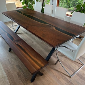 May include: A dark brown wooden dining table with a black epoxy river running down the center. The table has a black metal X-shaped base and is paired with a matching bench. The bench also has a black epoxy river running down the center and a black metal X-shaped base.