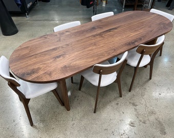 May include: A large oval wooden dining table with a natural wood finish. The table is surrounded by six white upholstered chairs with wooden legs.