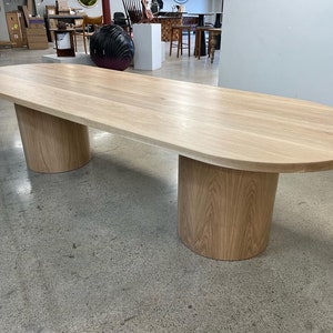 May include: A large oval wooden table with a light wood finish. The table has two cylindrical legs made of the same wood. The table is sitting on a concrete floor.