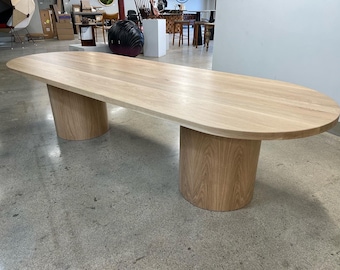May include: A large oval wooden table with a light wood finish. The table has two cylindrical legs made of the same wood. The table is sitting on a concrete floor.