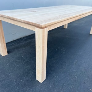 May include: A light wood dining table with a rectangular top and four square legs. The table is made of unfinished wood and has a natural grain pattern.