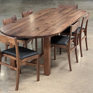 May include: A large oval walnut wood dining table with six chairs. The table has a natural wood finish and the chairs have black leather seats.