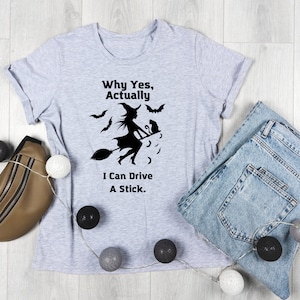 Può includere: Una maglietta grigia con una silhouette nera di una strega che vola su una scopa con un gatto nero. Il testo sulla maglietta recita "Why Yes, Actually, I Can Drive A Stick."
