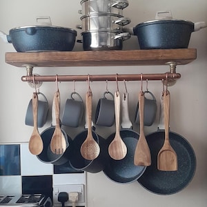 May include: A wooden shelf with copper pipe accents holds a collection of cookware.  The shelf has a black and grey speckled pot, a silver steamer, and a black and grey speckled frying pan.  The copper pipe is used to hang wooden spoons and spatulas.