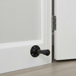 May include: Black industrial-style door stop mounted on a white baseboard. The door stop features a circular base with a short, cylindrical arm extending outwards. The door is partially open, revealing a hinge.