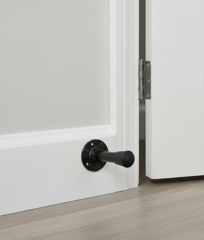 May include: Black industrial-style door stop mounted on a white baseboard. The door stop features a circular base with a short, cylindrical arm extending outwards. The door is partially open, revealing a hinge.