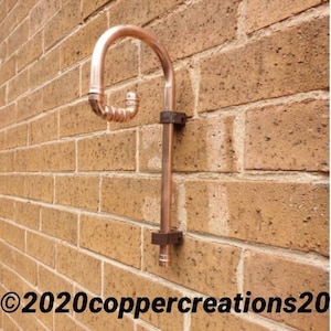 Può includere: Un raccordo per tubi in rame montato su un muro di mattoni. Il raccordo ha una parte superiore curva e una sezione di tubo verticale, tenuta in posizione da staffe metalliche. Il muro di mattoni è di un caldo colore terroso. Il testo "©2020coppercreations20" è visibile.