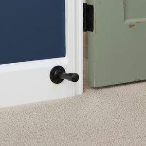 May include: A black door stop installed on a white door frame. The door stop is made of metal and has a cylindrical shape. A blue door is visible in the background, along with a green door.