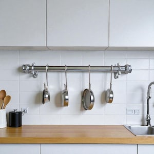 May include: A chrome pot rack with four stainless steel pans hanging in a kitchen. The rack is fixed to a white tiled wall above a wooden worktop. The pans are different sizes and styles.