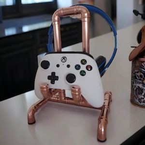 May include: Copper pipe gaming controller and headset stand. The stand holds a white Xbox controller with black buttons and a blue and black headset. The stand is made of copper pipes and fittings.