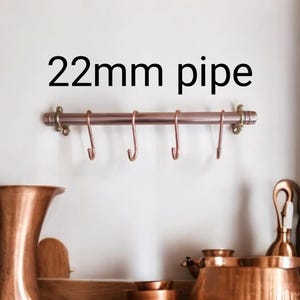 Può includere: Un portautensili da cucina in tubo di rame con cinque ganci. Il tubo ha un diametro di 22 mm e una finitura oro rosa. Il portautensili è montato su una parete bianca.