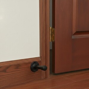 May include: A black door stop installed on a wooden baseboard. The door stop is made of metal and has a rounded base and a cylindrical handle. The door is a dark brown color and is slightly ajar, revealing a brass hinge.