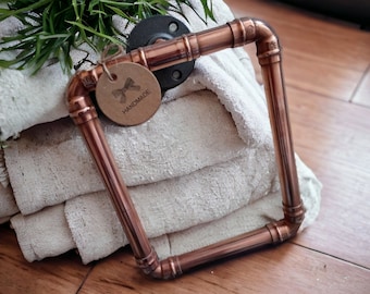 May include: A copper pipe frame with a handmade tag attached. The frame is sitting on top of a stack of white towels.