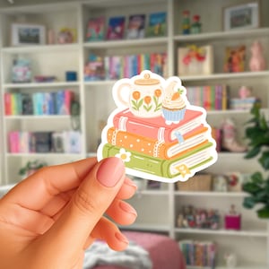 Könnte beinhalten: Ein gestanzter Aufkleber mit einem Stapel aus drei Büchern in Rosa, Orange und Grün. Eine weiße Teekanne mit orangefarbenen Tulpen und ein kleiner Cupcake mit einer Karotte als Topping stehen auf den Büchern. Der Aufkleber hat einen weißen Rand.