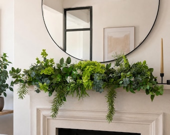 Op de afbeelding: Een weelderige kunstgroenslinger met een mix van levendig limoengroen, diep bosgroen en saliekleurig blad. Het arrangement bevat diverse bladvormen, delicate witte bloesems en naar beneden hangende stengels op een schoorsteenmantel.