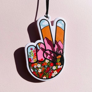 May include: Peace sign car air freshener with a colorful mountain and flower design. The peace sign is pink and orange with a green, blue, and white mountain scene inside. The bottom of the peace sign is filled with colorful flowers.