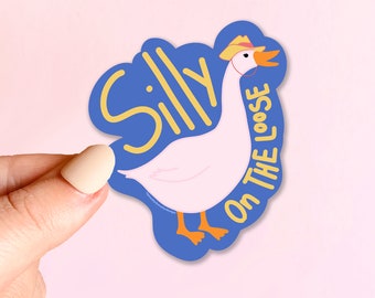 Puede incluir: Una pegatina troquelada con un ganso de dibujos animados con un sombrero de vaquero. El ganso es blanco con patas y pico naranjas, sobre un fondo azul con las palabras "Silly On The Loose" en amarillo.