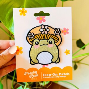 May include: Iron-on patch featuring a cartoon frog wearing a mushroom hat with pink flowers. The patch is on a white card with the text "Peachy Keen Iron-On Patch @peachykeenbydesign".