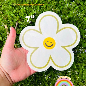 Op de afbeelding: Een witte daisy sticker van 17,8 cm met een geel smiley gezicht in het midden. De sticker is op een groene achtergrond.