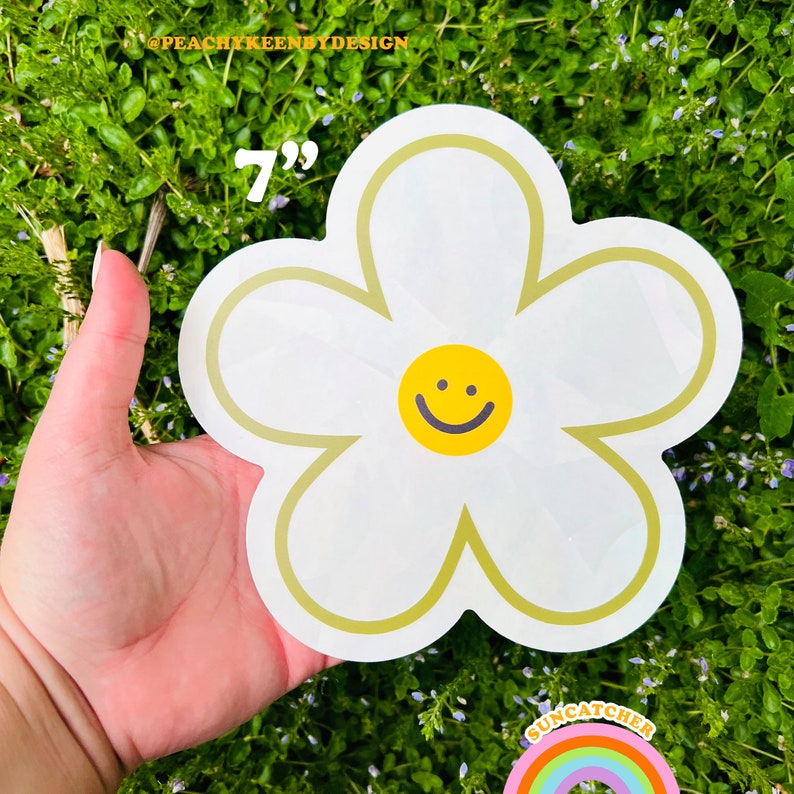 Op de afbeelding: Een witte daisy sticker van 17,8 cm met een geel smiley gezicht in het midden. De sticker is op een groene achtergrond.