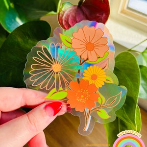 Può includere: Un adesivo olografico con un bouquet di fiori in tonalità arancio, giallo e blu. I fiori sono contornati di nero e hanno uno stile retrò. L'adesivo è su uno sfondo trasparente e ha una finitura arcobaleno iridescente.