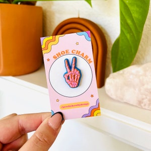 May include: Pink skeleton hand peace sign shoe charm with blue outline. The charm is on a pink card with the text "Shoe Charm" and "#peachykeenbydesign".