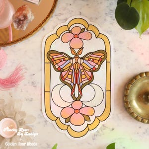 May include: A sticker with a stained-glass design featuring a moth with colorful wings, set within a yellow and white frame. Two pink flowers are at the top and bottom. The text "Peachy Keen By Design + Golden Hour Abode" is at the bottom.