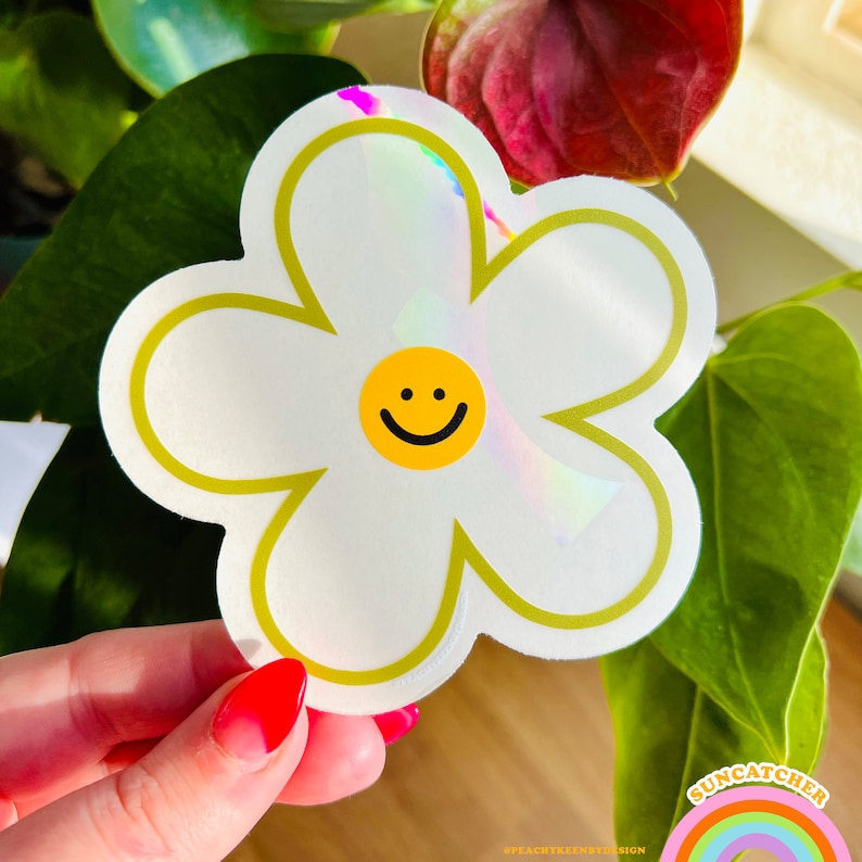 Op de afbeelding: Een witte madeliefjesticker met een geel smiley-gezicht in het midden. De sticker heeft een holografische afwerking en een gouden omtrek.