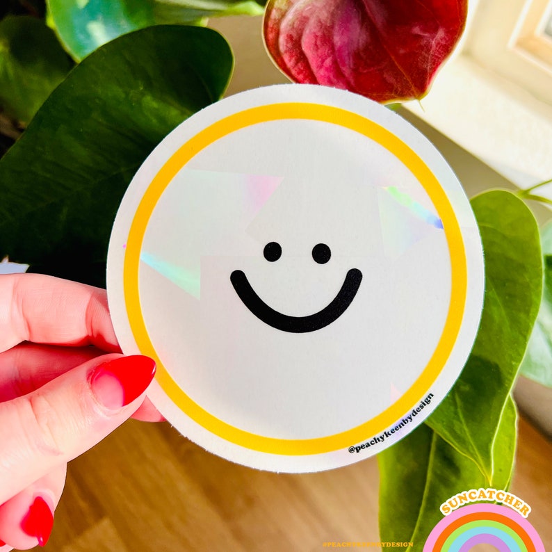Pu&ograve; includere: Un adesivo bianco con un bordo giallo e una faccina sorridente nera. L'adesivo ha una finitura olografica e il testo "@peachykeenbydesign" &egrave; stampato in basso.
