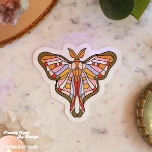May include: A colorful moth sticker with a stained-glass effect. The sticker features a symmetrical design with orange, pink, yellow, and purple hues, outlined in brown. The text "Peachy Keen By Design + Golden Hour Abode" is visible.