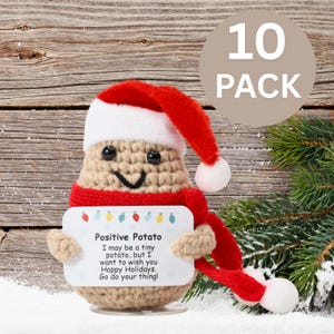 May include: A crocheted potato wearing a red Santa hat and scarf holds a card that reads "Positive Potato I may be a tiny potato, but I want to wish you Happy Holidays. Go do your thing!" The potato is sitting on a white surface with a snowy background.