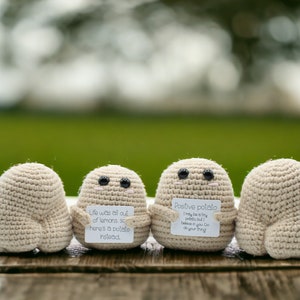 May include: Four crocheted potato-shaped figures with black button eyes are arranged on a wooden surface. Two figures hold small signs with text. One sign reads "Life was all out of lemons so here's a potato instead." The other says "Positive potato."