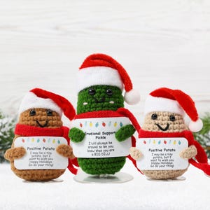 May include: Three crocheted Christmas ornaments, a green pickle, and two brown potatoes, each wearing a red Santa hat and holding a sign with a holiday message. The pickle's sign reads "Emotional Support Pickle I will always be around to let you know that you are a BIG DILL". The potatoes' signs read "Positive Potato I may be a tiny potato, but I want to wish you Happy Holidays. Go do your thing!"